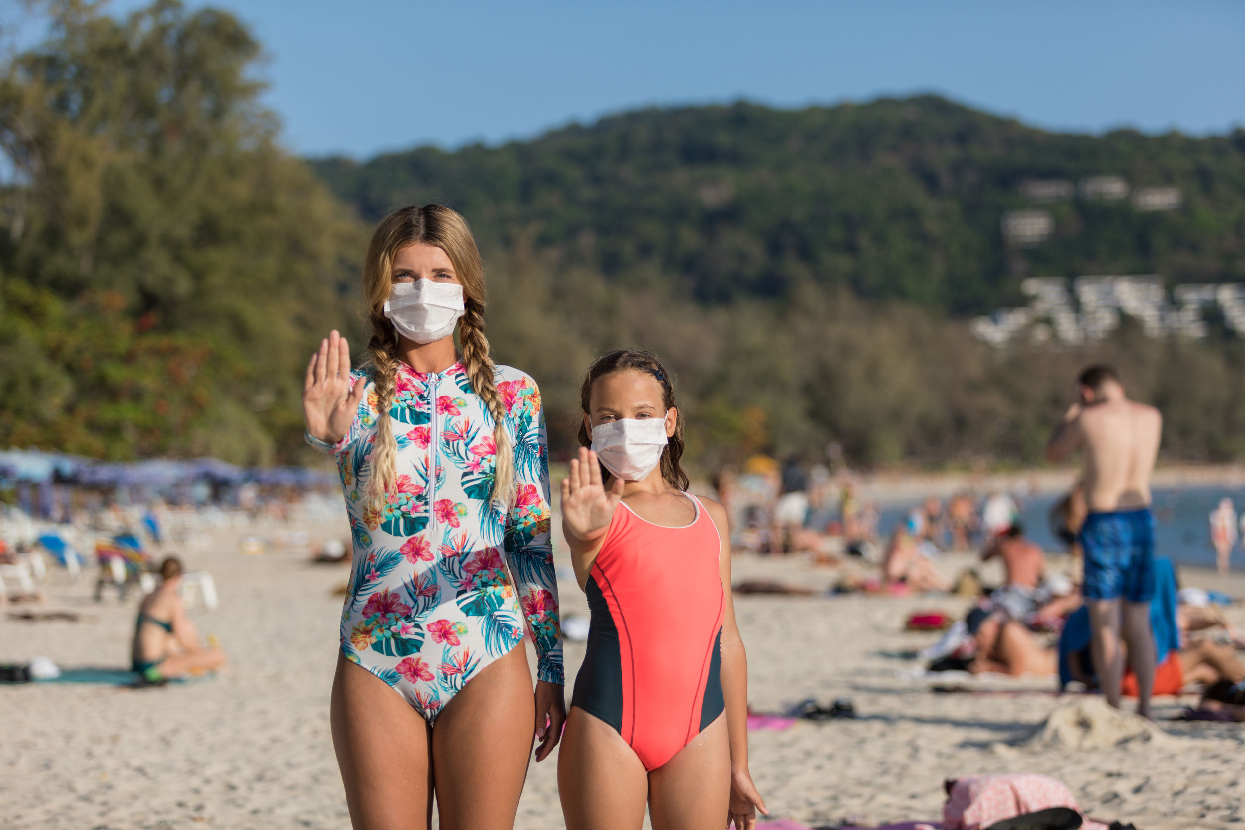 A woman and a child in a protective surgical mask on their face are standing on the beach in swimsuits. Chinese coronavirus disease COVID-19 is dangerous